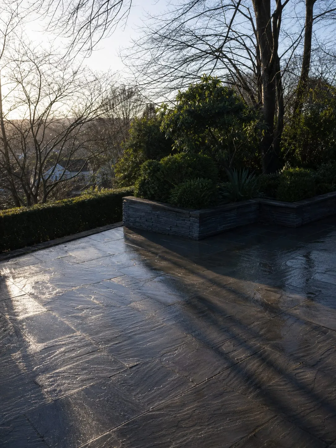 An upper terrace paved in riven Welsh slate, wet and dark, with low winter sun raking long shadows across it.