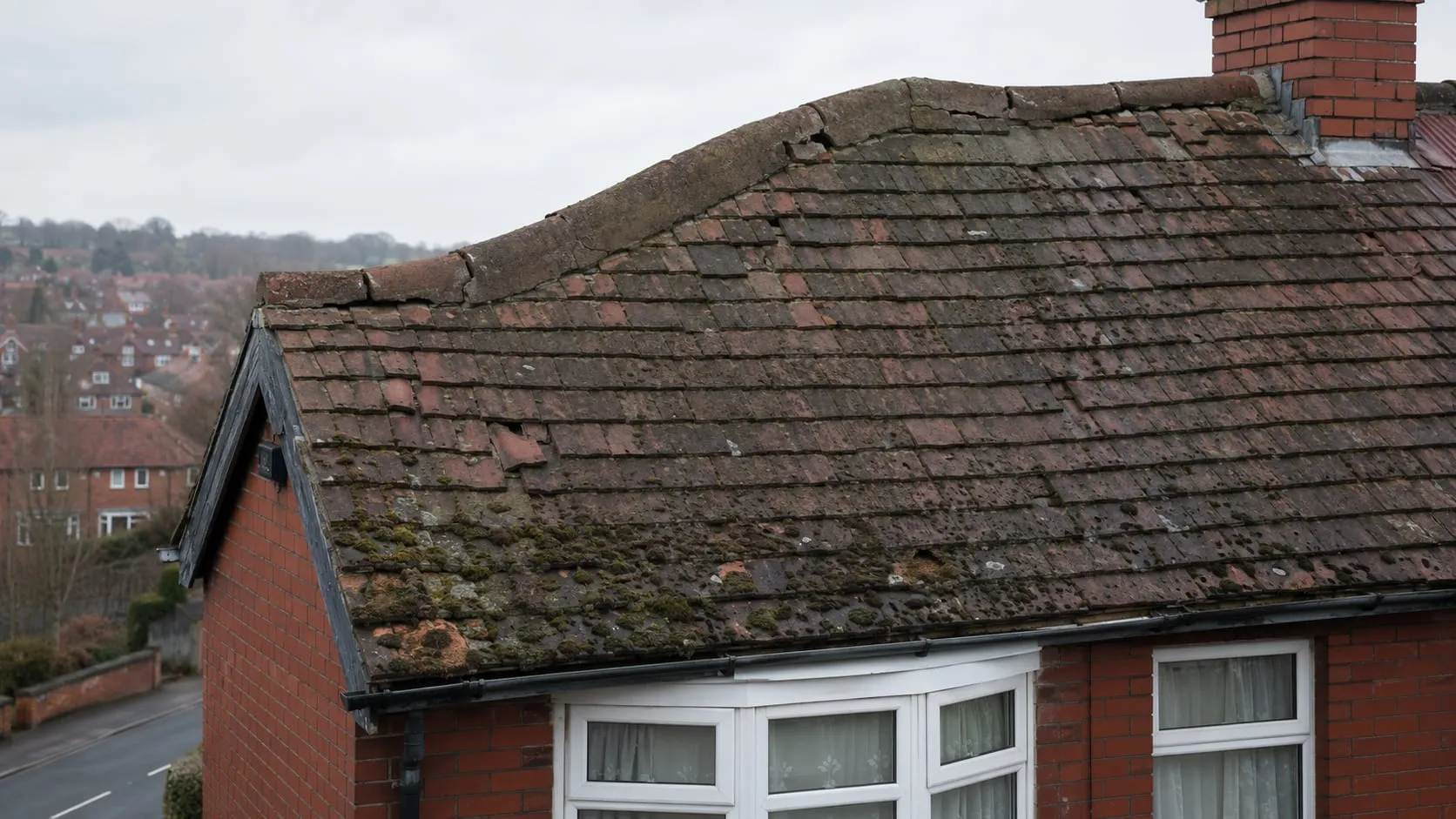 A tired clay plain-tiled roof on a 1930s semi before work: faded blotchy tiles, cracked and slipped units, heavy moss and a dipped roof line.